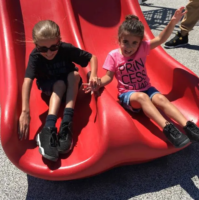 Two kids go down a red slide while holding hands.