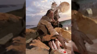 A grieving person kneels by a lakeside grave, while another person holds a large drum behind them.
