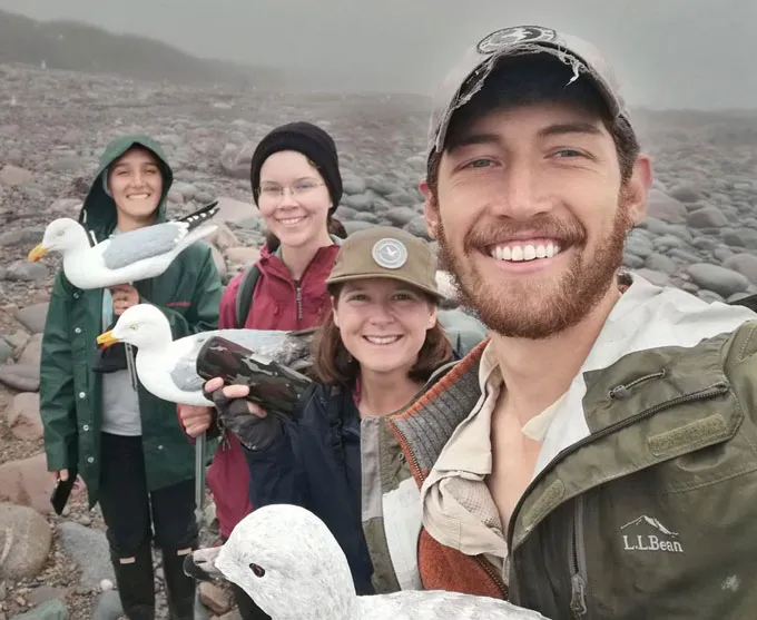 Four people are grinning on a rocky foggy cold-looking beach. They are holding plastic seagulls.