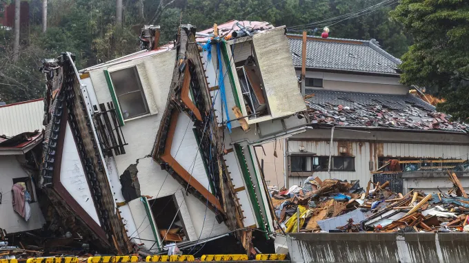 A house partially collapsed and tilted sharply onto its side amid scattered debris and other damaged buildings.