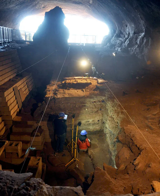 A helmeted researcher stands in a massive pit dug into the floor of a cave, surrounded by archaeological equipment