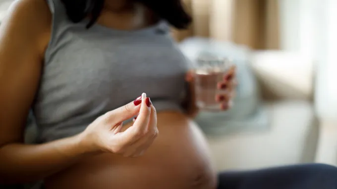 A pregnant person sits on a couch holding a glass of water in one hand and a pill in the other