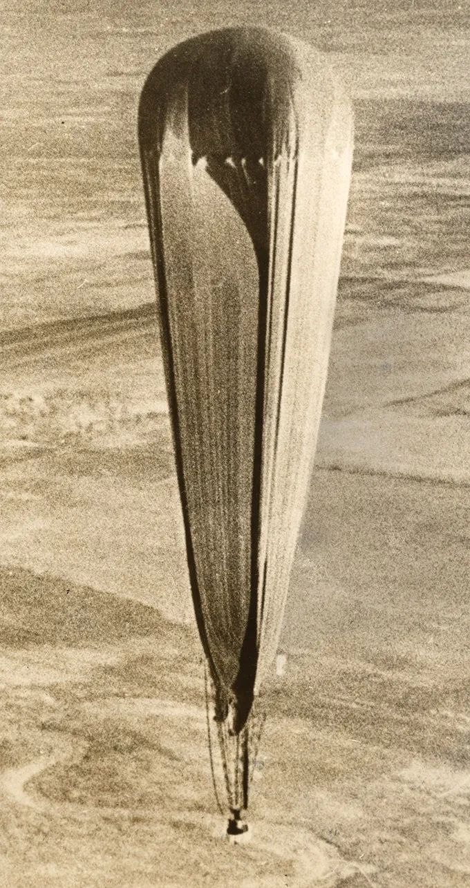 An aerial view of an early high-altitude balloon called The Explorer II balloon mission, with a long, tapered shape drifting above the landscape.