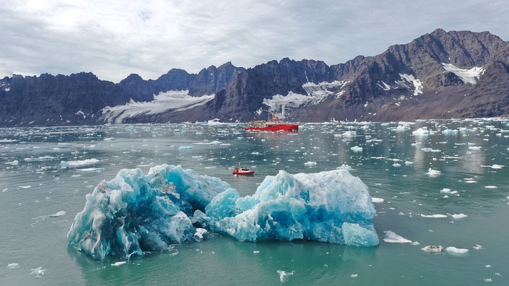 Two boats float in icy waters. Rocky cliffs loom in the background, while a big chunk of white and blue ice is in the foreground. It dwarfs the smaller of two red boats.