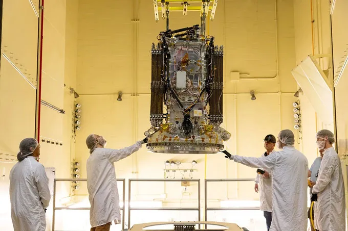 Engineers in white coats and gloves examine a robotic spacecraft suspended from above in a white-walled room.