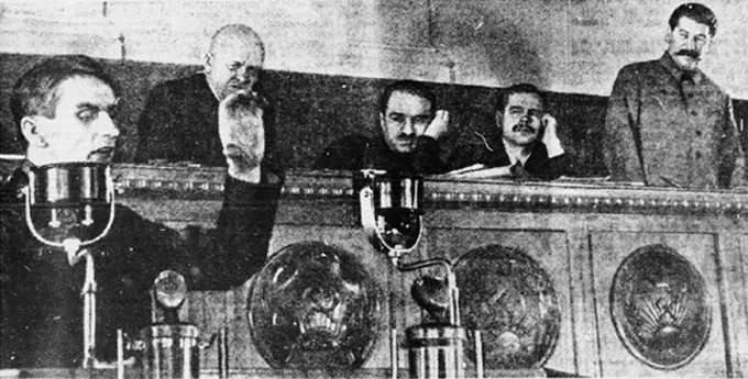 A black and white historical photo showing Trofim Lysenko giving a speech. Lysenko's chin is obscured by a lamp. He has his left hand raised. His fingers are bent and thumb extended as if indicating how large something is. A raised bench is behind him. Lysenko's hand blocks the chin of a bald man, Stanislav Kosior, who is partially standing. Two men, Anastas Mikoyan and Andrei Andreyev, with dark hair and mustaches sit and rest their heads in their left hands and while watching Lysenko. Joseph Stalin stands behind the bench at the far right of the photo.