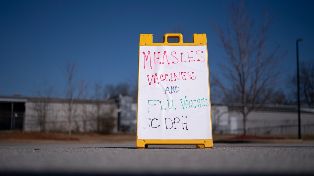 A folding sign that says "Measles vaccines and flu vaccines SC DPH" stands in the middle of a sidewalk with a distant building in the background