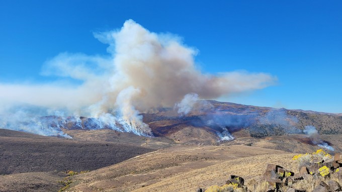 Smoke and flame rise along a ridge of dry, rolling hills under a blue sky.