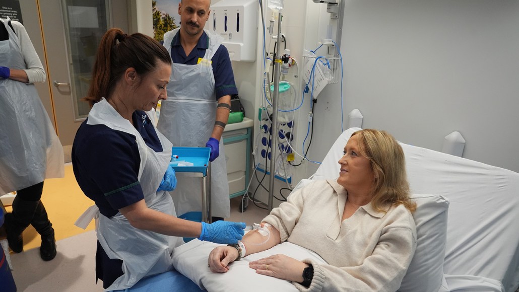 A person sits in a hospital bed while receiving treatment from a medical provider through an infusion in their arm