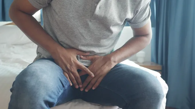 A man in a gray shirt and jeans clasps both hands over his groin area.