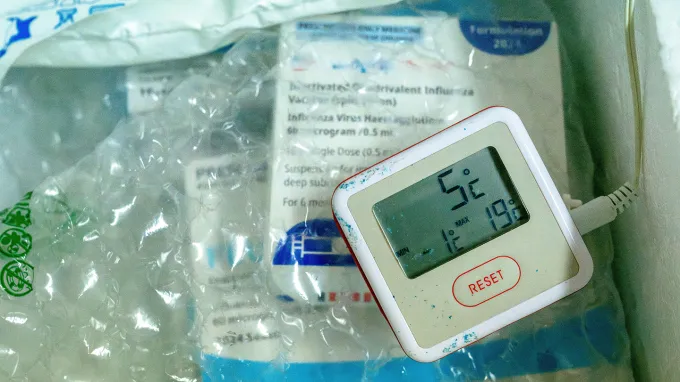 A thermometer layered over bubble wrap and vaccine packaging in a cooler reads 5 degrees Celsius