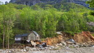 A partially collapsed home sits on the side of a caved-in riverbed. Bright green dense forest is behind it, with a mountain popping up in the background against the sky.