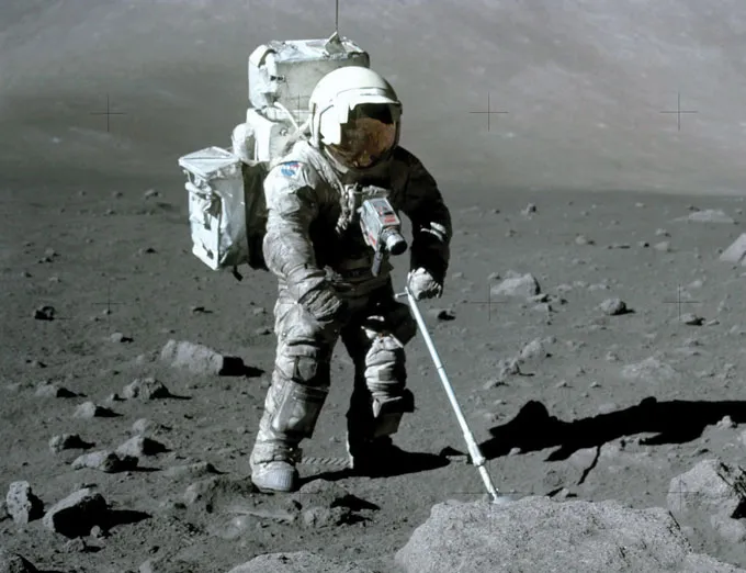 An astronaut in a bulky spacesuit stands on the rocky surface of the Moon, using a handheld tool to collect a sample. The gray, dusty landscape stretches into the distance, with scattered rocks and the stark horizon.