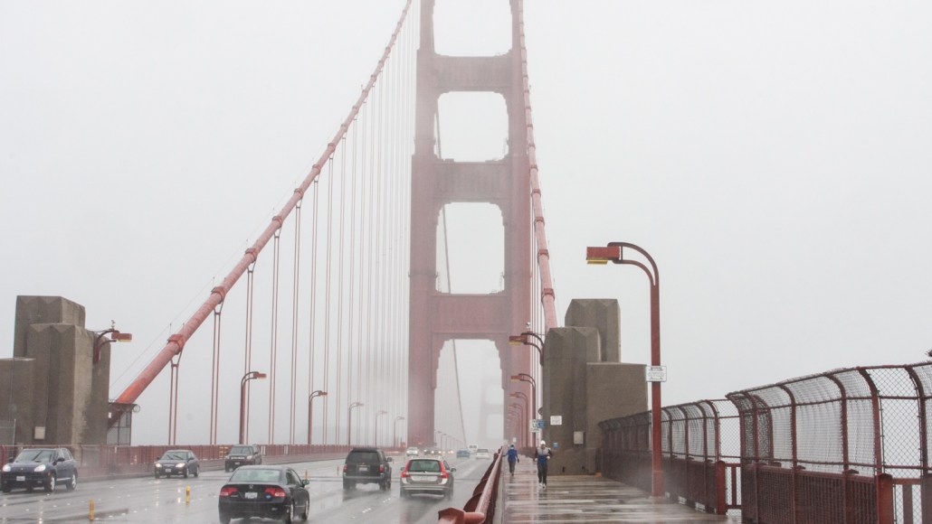 Vehicles and pedestrians cross the Golden Gate Bridge on a foggy damp-looking day.