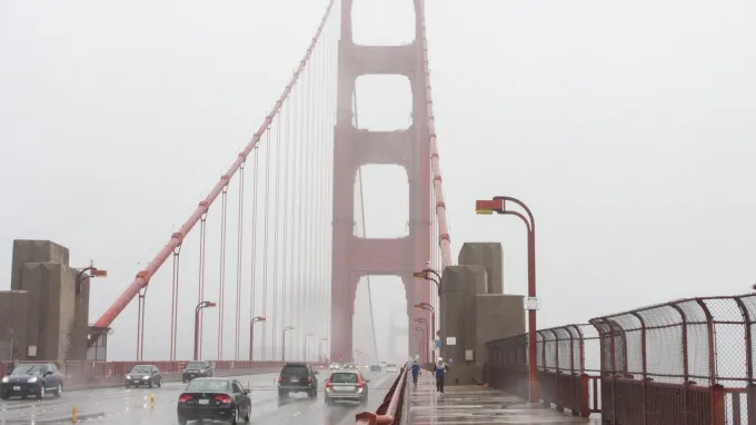 Vehicles and pedestrians cross the Golden Gate Bridge on a foggy damp-looking day.