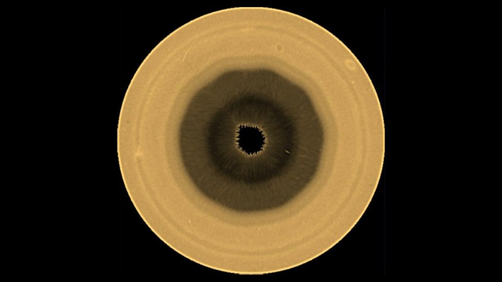 A golden colored orb has a very dark center, surrounded by lightening colors as you move outward. It shows a weird weather pattern at Saturn's south pole.