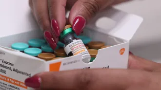 A person with red nails picks up a bottle of shingles vaccine out of a box.
