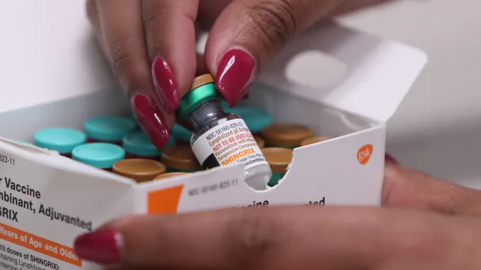A person with red nails picks up a bottle of shingles vaccine out of a box.